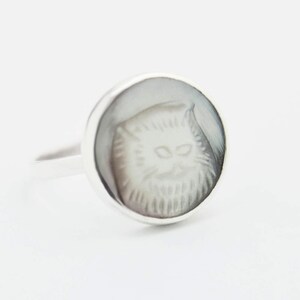 May include: A silver ring featuring a white cat cameo. The cameo is set in a round bezel and the ring band is plain.