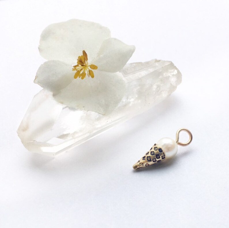 May include: Gold charm shaped like an ice cream cone with a white pearl on top. The charm is sitting on a clear quartz crystal.