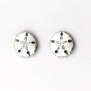 May include: A pair of silver sand dollar stud earrings. The earrings are small and round, with a detailed sand dollar design.