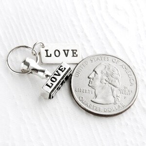 May include: A silver charm with a small stamp that reads "LOVE" and a tag that reads "LOVE". The charm is next to a US quarter.