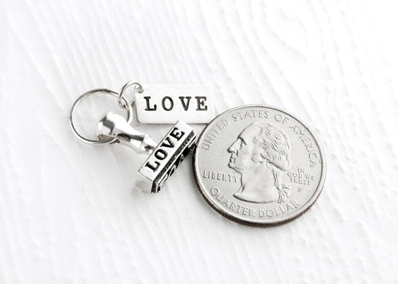May include: A silver charm with a small stamp that reads "LOVE" and a tag that reads "LOVE". The charm is next to a US quarter.