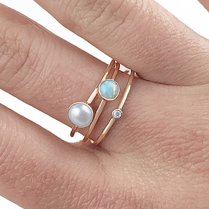 Puede incluir: Tres anillos de oro con diferentes piedras preciosas. El anillo superior tiene un ópalo azul claro, el anillo del medio tiene una perla blanca y el anillo inferior tiene un pequeño diamante transparente.