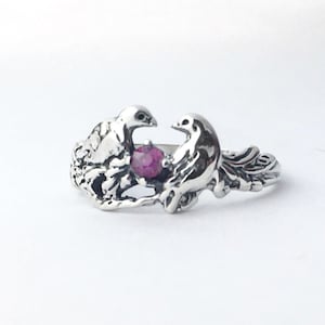 May include: A silver ring featuring two doves facing each other, with a small, round, red gemstone between them. The ring has a detailed, sculpted design, and the doves are perched on a branch.