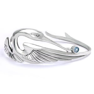 Könnte beinhalten: Ein silberfarbenes Armband mit einem stilisierten Vogeldesign. Das Armband zeigt den Kopf, die Flügel und den Schwanz eines Vogels, mit einem kleinen, runden, hellblauen Edelstein in der Nähe des Schwanzes. Auf weißem Hintergrund.
