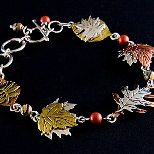 May include: A silver bracelet with a toggle clasp features five leaf charms made of silver, copper, and gold. The leaves are layered and have a textured finish. The bracelet also has red and brown beads.