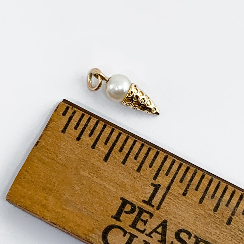 May include: Gold charm in the shape of an ice cream cone with a white pearl on top. The charm is about 1 inch long.