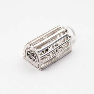 Puede incluir: Un charm de plata con forma de jaula. La jaula está hecha de finas barras de metal y tiene una parte superior redondeada. El charm está diseñado para ser fijado a una pulsera o collar.