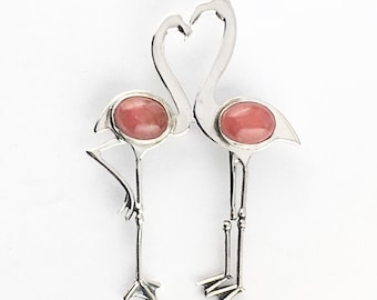 May include: A silver flamingo brooch featuring two stylized flamingos with their necks forming a heart shape. Each flamingo has a pink oval-shaped stone set in its body. The brooch is set against a white background.
