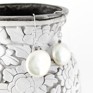 May include: A pair of silver dangle earrings with white pearl-like round drops. The earrings are displayed on a white ceramic vase with floral designs.