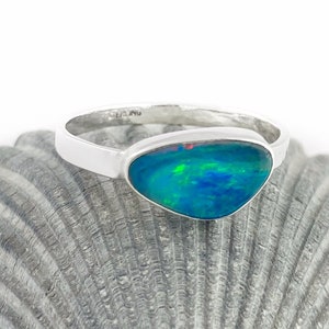 May include: A silver ring with a blue, green, and red opal gemstone. The ring is set on a seashell background.