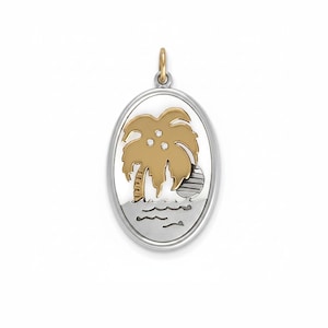 May include: An oval pendant featuring a two-tone metal design with a gold-colored palm tree silhouette set against a silver-colored background. The palm tree includes three small embedded clear stones, and the base depicts stylized silver waves.