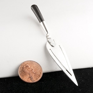 May include: A silver bookmark with a dark wooden handle. The bookmark has a long, pointed design with two vertical cutouts. A U.S. penny is next to the bookmark for size comparison.