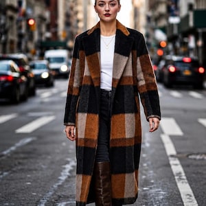 May include: A long, knee-length coat featuring a large, bold plaid pattern in shades of brown, black, and tan. The coat has a wide lapel collar, long sleeves, and a straight-cut silhouette. It is worn over a plain white t-shirt and dark trousers.