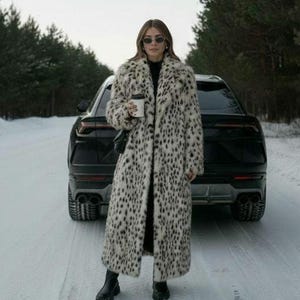 May include: A long, faux fur coat with a black and white spotted pattern. The coat is worn over a black outfit, paired with black boots. The person is holding a coffee cup and standing on a snow-covered road with a black SUV in the background.