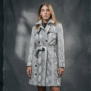 May include: A double-breasted trench coat featuring a grey and white snakeskin print pattern, a wide notched collar, long sleeves with buckled cuffs, and a matching tie belt at the waist, paired with black knee-high leather boots.