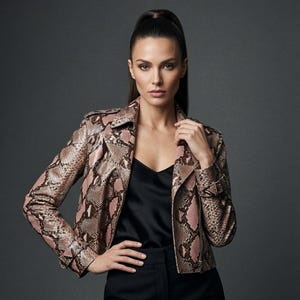 May include: A moto-style jacket featuring a pink and brown snakeskin pattern, a notched collar, long sleeves with buckled cuffs, and a front zipper closure. The jacket is worn over a black camisole and black trousers against a dark gray background.
