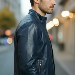 May include: A navy blue leather jacket with a stand-up collar and a zippered chest pocket. The jacket has a tailored fit and is worn by a person in an outdoor setting. The jacket is a classic style.
