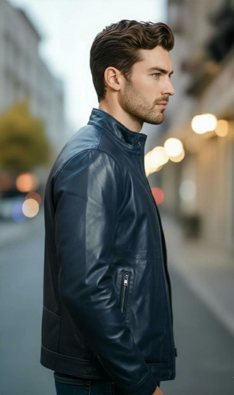 May include: A navy blue leather jacket with a stand-up collar and a zippered chest pocket. The jacket has a tailored fit and is worn by a person in an outdoor setting. The jacket is a classic style.