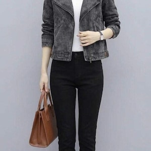 May include: A woman wearing a dark gray denim jacket with a white shirt underneath and black pants. She is carrying a brown leather handbag and wearing black boots. The jacket has a classic asymmetrical zipper design.