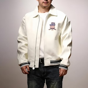 May include: Cream-colored jacket with a black zipper and a black and white striped hem. The jacket features a crest with the text "JIREX" and a design resembling the American flag. The jacket is worn with a white shirt and blue jeans.