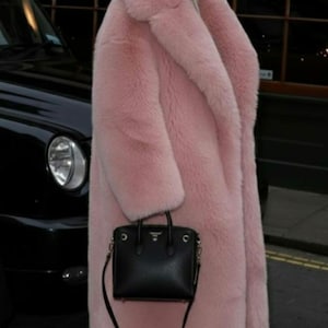 May include: A long, light pink faux fur coat with a wide collar. A black handbag with a shoulder strap is held in front of the coat. Black high-heeled sandals complete the look. The coat is a statement piece.
