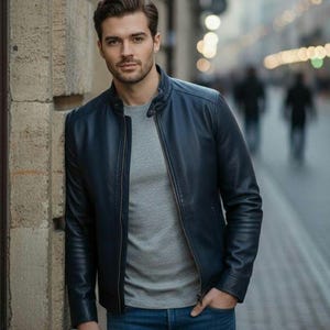 May include: A navy blue leather jacket with a zip-up front, worn over a gray crew neck shirt and blue jeans. The jacket has a classic, streamlined design. The person is standing outdoors.