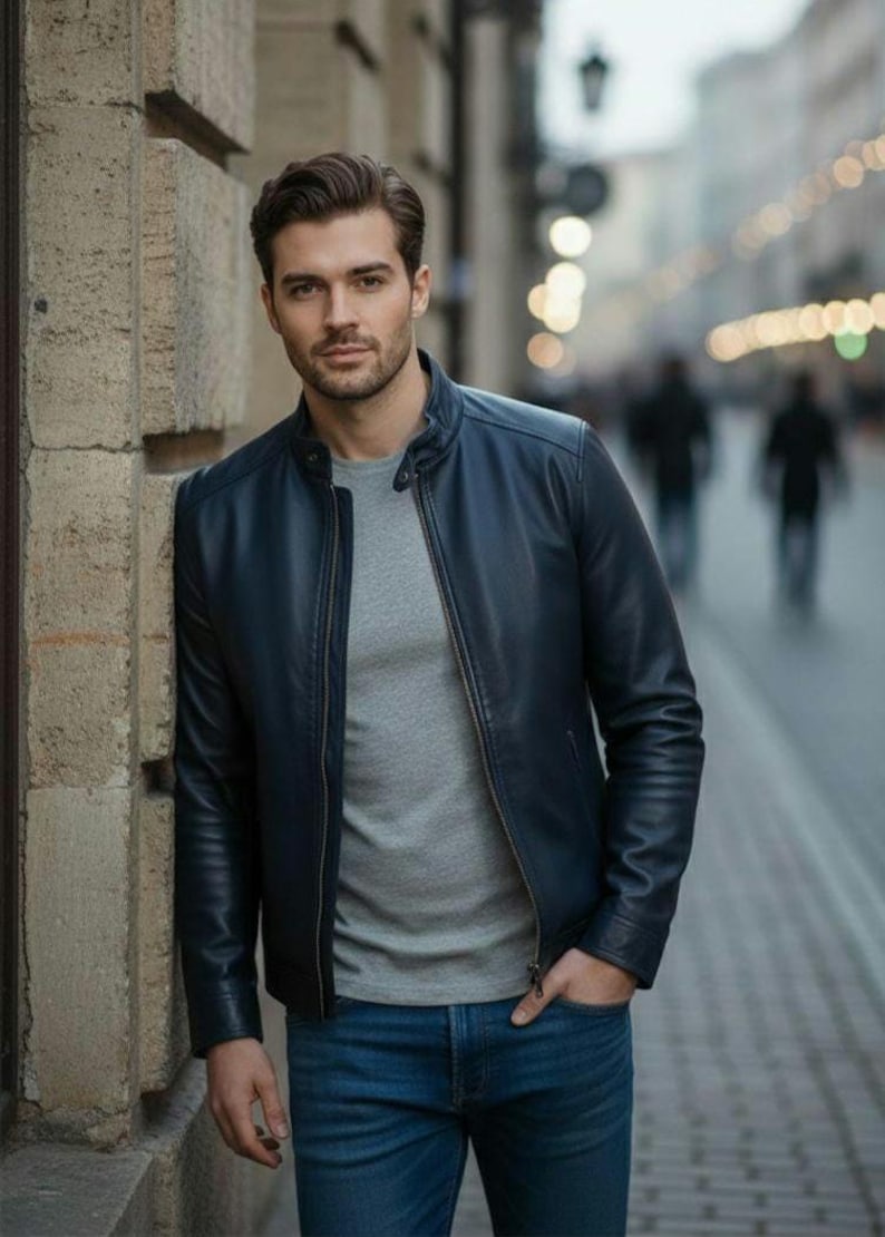 May include: A navy blue leather jacket with a zip-up front, worn over a gray crew neck shirt and blue jeans. The jacket has a classic, streamlined design. The person is standing outdoors.