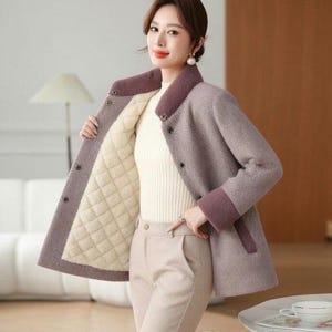 May include: A mauve herringbone-patterned coat with a quilted cream-colored lining. The coat features a dark purple collar and cuffs, and is buttoned up. The model is wearing a cream-colored sweater and tan pants. The text "Royal Apparel" is at the bottom.