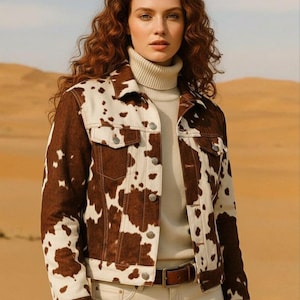 May include: A brown and white cow print denim jacket with silver buttons. The jacket is worn over a cream-colored turtleneck and paired with light-colored jeans and a brown belt. The background is a desert landscape.