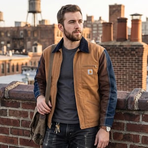 May include: A tan jacket with a dark blue corduroy collar and dark blue sleeves, worn over a grey t-shirt. The jacket features a small Carhartt logo patch on the left chest pocket. A brown messenger bag strap crosses the chest, and keys hang from the belt loop of dark denim jeans.