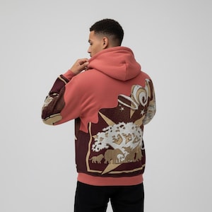 May include: A coral-colored hoodie with a detailed tapestry design on the back. The tapestry features a tree, animals, and abstract shapes in shades of brown, gold, and white. The sleeves have a patterned design in similar colors.