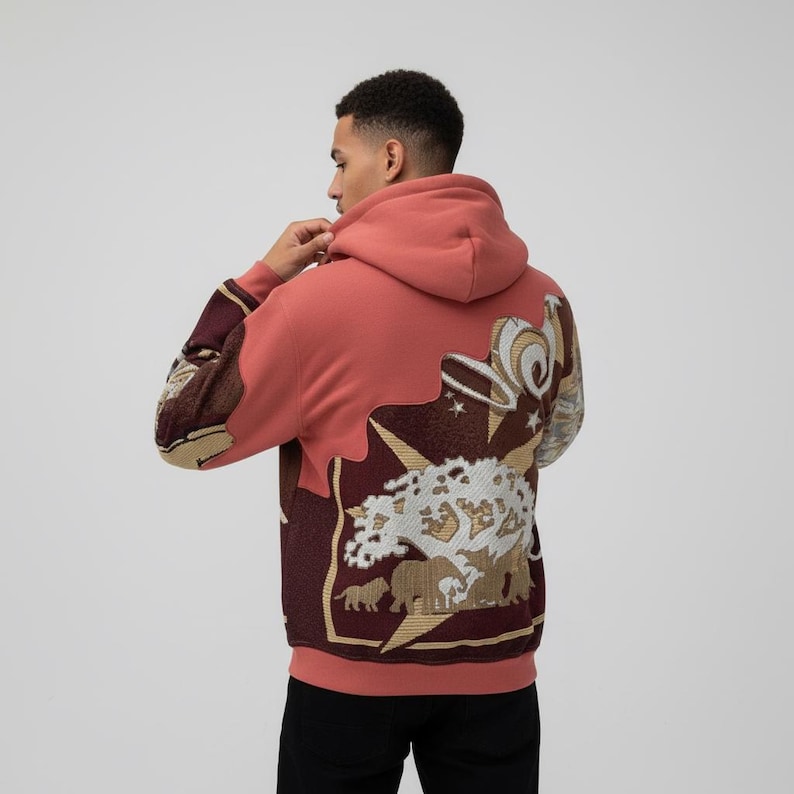 May include: A coral-colored hoodie with a detailed tapestry design on the back. The tapestry features a tree, animals, and abstract shapes in shades of brown, gold, and white. The sleeves have a patterned design in similar colors.