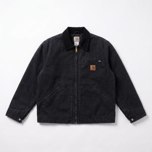 May include: A black canvas work jacket featuring a corduroy collar, a full-length front zip, and long sleeves with snap-button cuffs. The jacket includes a zipped chest pocket, two lower front pockets, and a Carhartt brand patch on the chest.