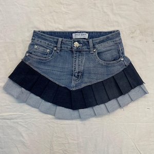 以下が含まれることがあります： Wax Jean Collectionのラベル、金属製のボタン留め、銀色のリベットが付いた2つのフロントポケットを備えた青いデニムのミニスカート。スカートは、ダークネイビーブルーの中段とライトブルーの下段の層状のプリーツヘムデザインが特徴です。