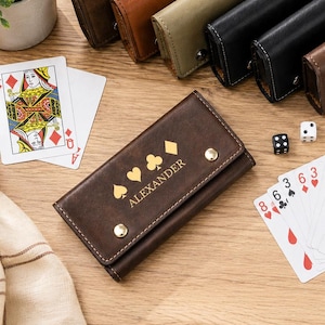 May include: A collection of rectangular leather cases with snap closures in various colors including brown, tan, and black. One case features the name ALEXANDER printed in gold with card suit symbols. Playing cards, dice, and a pen are also present.