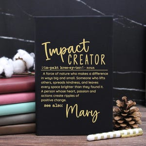 May include: A black notebook with gold text that reads Impact Creator, a definition of the term, and the name Mary. The notebook is positioned next to a stack of colorful journals, a pinecone, and a white and gold striped pen.