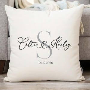 Puede incluir: Cojín cuadrado color crema con texto personalizado. El cojín presenta los nombres "Colten & Hailey" en una fuente cursiva, con una gran "S" gris en el centro y la fecha "01.12.2026" debajo. El cojín está sobre un sofá gris.