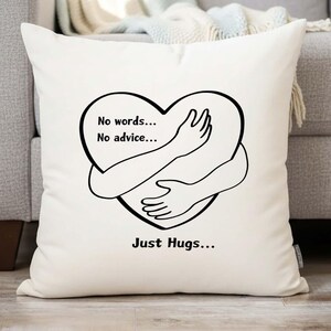 Puede incluir: Cojín crema con un diseño negro en forma de corazón con brazos abrazándose. El texto en el cojín dice "No words... No advice... Just Hugs...". El cojín está sobre un sofá gris.