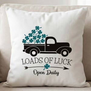 Puede incluir: Cojín cuadrado blanco con un dibujo negro de un camión vintage lleno de tréboles verde azulado. El texto "LOADS OF LUCK" está encima de una flecha con un trébol, y "Open Daily" debajo. El cojín está sobre un sofá beige.