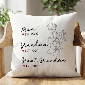 May include: Cream cushion with black text: "Mom EST 1960", "Grandma EST 1990", and "Great Grandma EST 2026". A floral design is on the right. The cushion is on a wooden chair, with plants in the background.