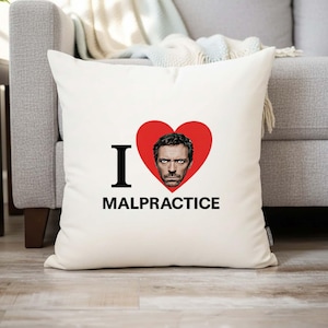 Puede incluir: Cojín cuadrado blanco con el texto negro "I MALPRACTICE". Un corazón rojo con el retrato de un hombre está entre la "I" y la palabra. El cojín está sobre un sofá gris.