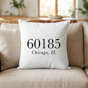 Könnte beinhalten: Weißes quadratisches Kissen mit dem schwarzen Text "60185 Chicago, IL". Das Kissen liegt auf einem beigen Sofa. Der Hintergrund umfasst einen Holztisch und Stühle.
