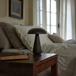 Puede incluir: Una lámpara de mesa negra con forma de hongo, con una parte superior redondeada y una base cónica. La lámpara está sobre una mesita de noche de madera oscura, junto a un libro y unas gafas. El fondo muestra una cama con ropa de cama blanca y una ventana.