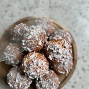 Pode incluir: Um close-up de um prato de madeira com uma pilha de pequenos doces redondos e marrons cobertos com açúcar em pó branco e granulado branco.