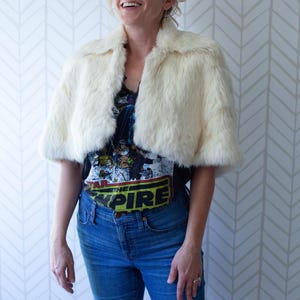 May include: Cream-colored faux fur cape with a wide collar. The cape is worn over a black graphic t-shirt and blue jeans. The t-shirt features the text "THE EMPIRE" in yellow and green.