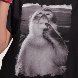 May include: Black t-shirt featuring a black and white image of a monkey in a thoughtful pose, with its finger touching its lip. The monkey is set against a blurred outdoor background. The shirt is made of a soft material.