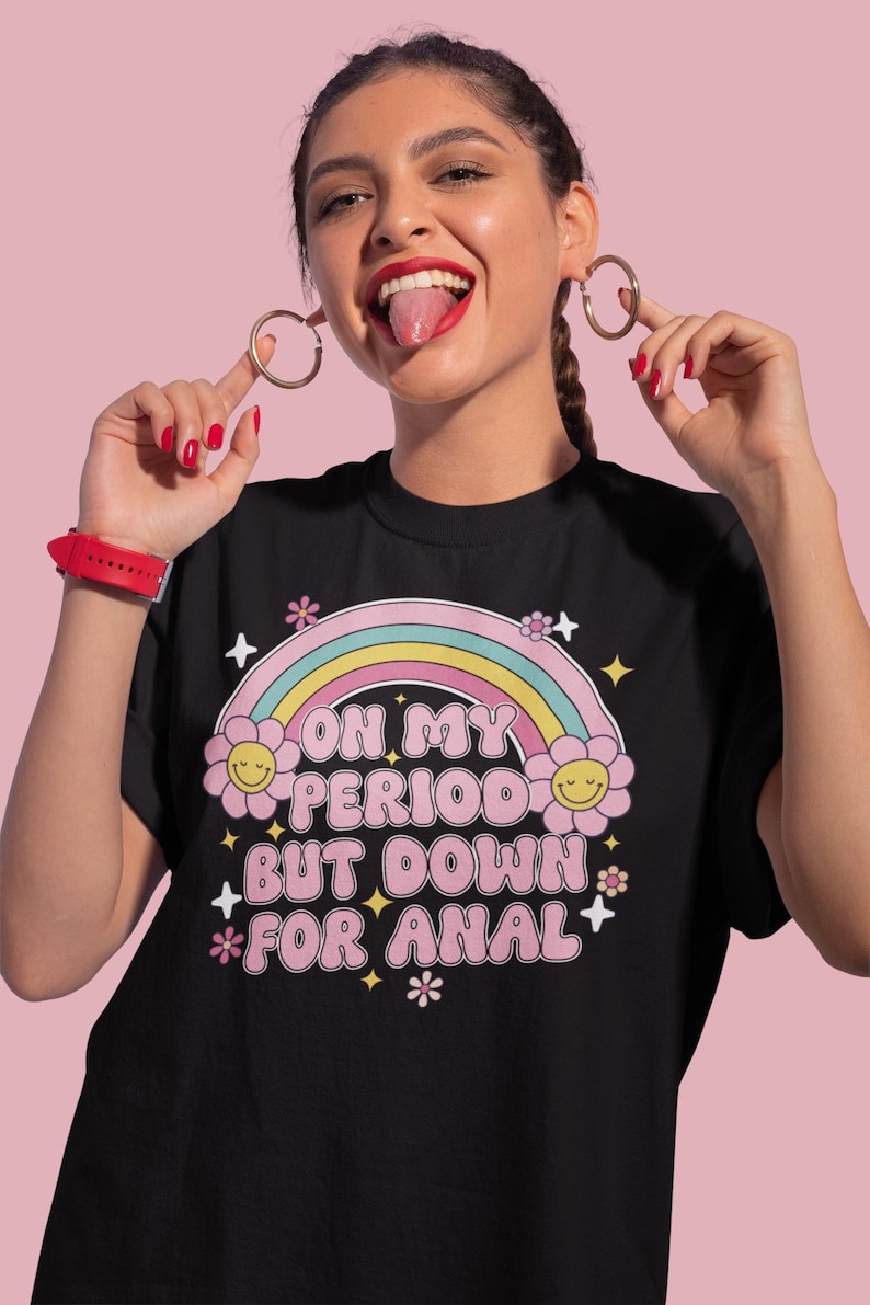 Puede incluir: Camiseta negra con un arco&iacute;ris y un gr&aacute;fico de flores. El texto de la camiseta dice "ON MY PERIOD BUT DOWN FOR ANAL". El dise&ntilde;o incluye colores amarillo, rosa y blanco.