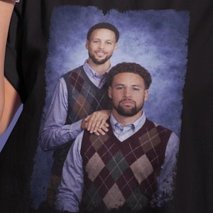 Peut inclure: T-shirt noir avec un portrait de deux hommes portant des gilets en losanges sur des chemises bleu clair. L'image a un effet vintage légèrement flou, avec un fond bleu.