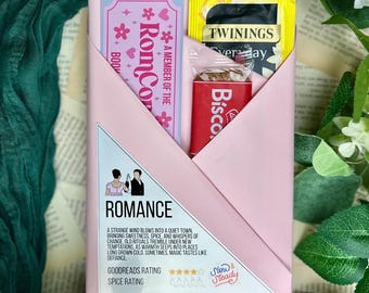 May include: A pink envelope-style package containing a bookmark, a tea bag, and a biscuit. The bookmark reads "RomCor" and "A Member of the Boo". The package is surrounded by greenery and flowers, with the word "Romance" visible.