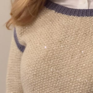 Può includere: Un maglione di lana color crema con un bordo viola intorno al collo e una striscia viola sulle maniche. Il maglione ha una leggera lucentezza su tutta la superficie.